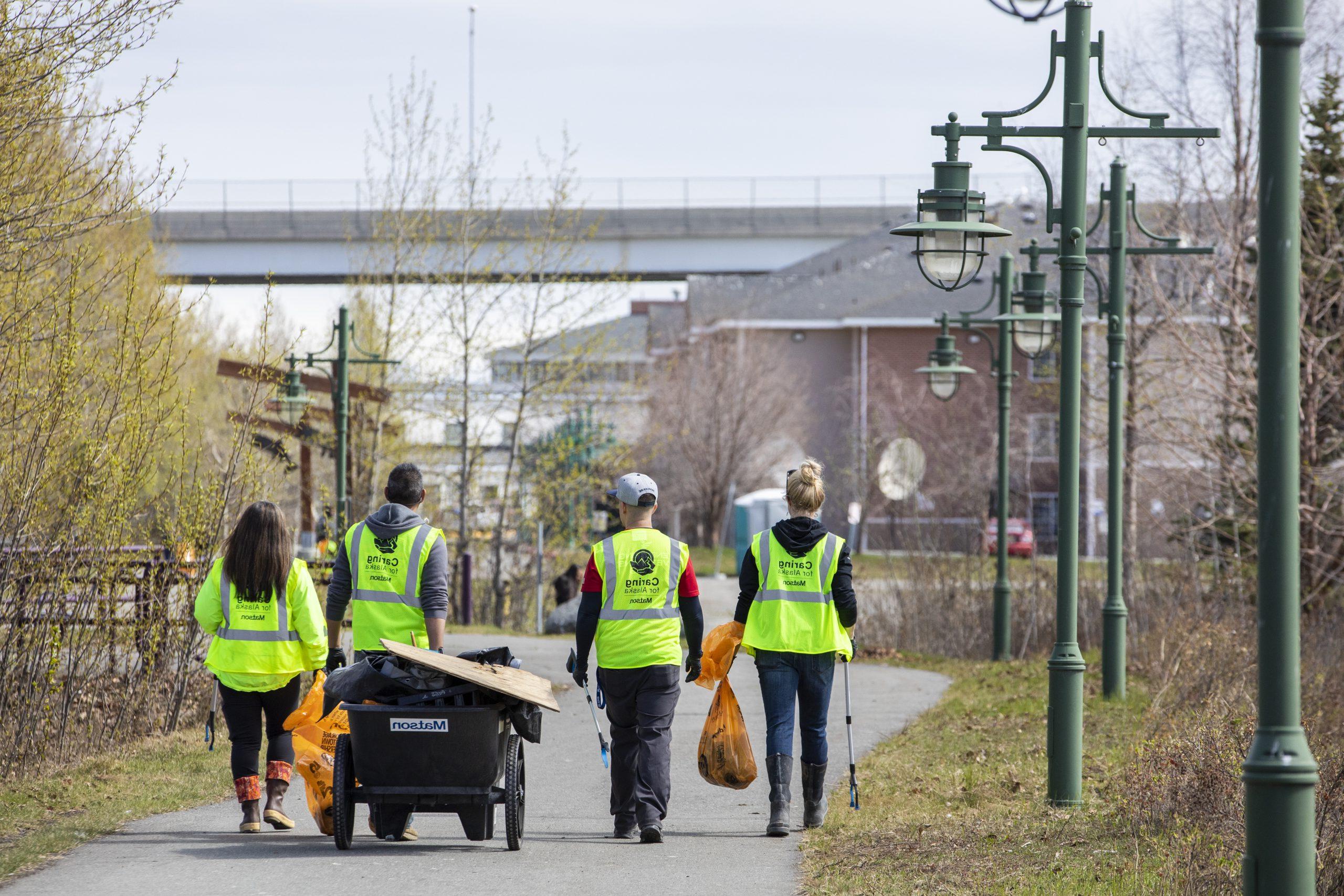 Volunteers wearing yellow safety vests walk along Ship Creek with a cart and orange bags full of trash.