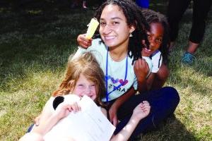 Two smiling young female campers at Hearts to Arts camp cozy up to a counselor on the grass.