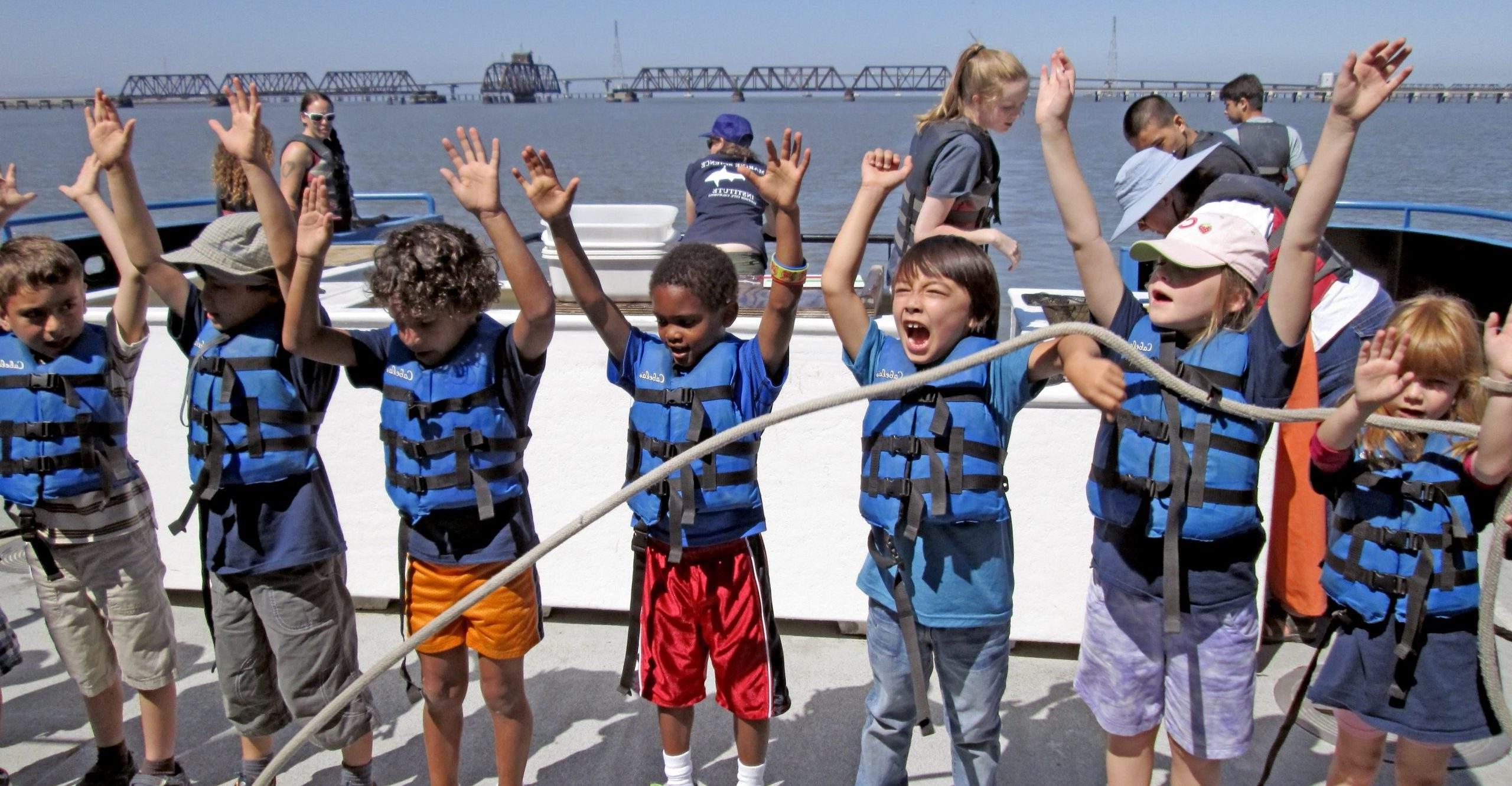 Children wearing blue life vests raise their arms on the deck of a boat.