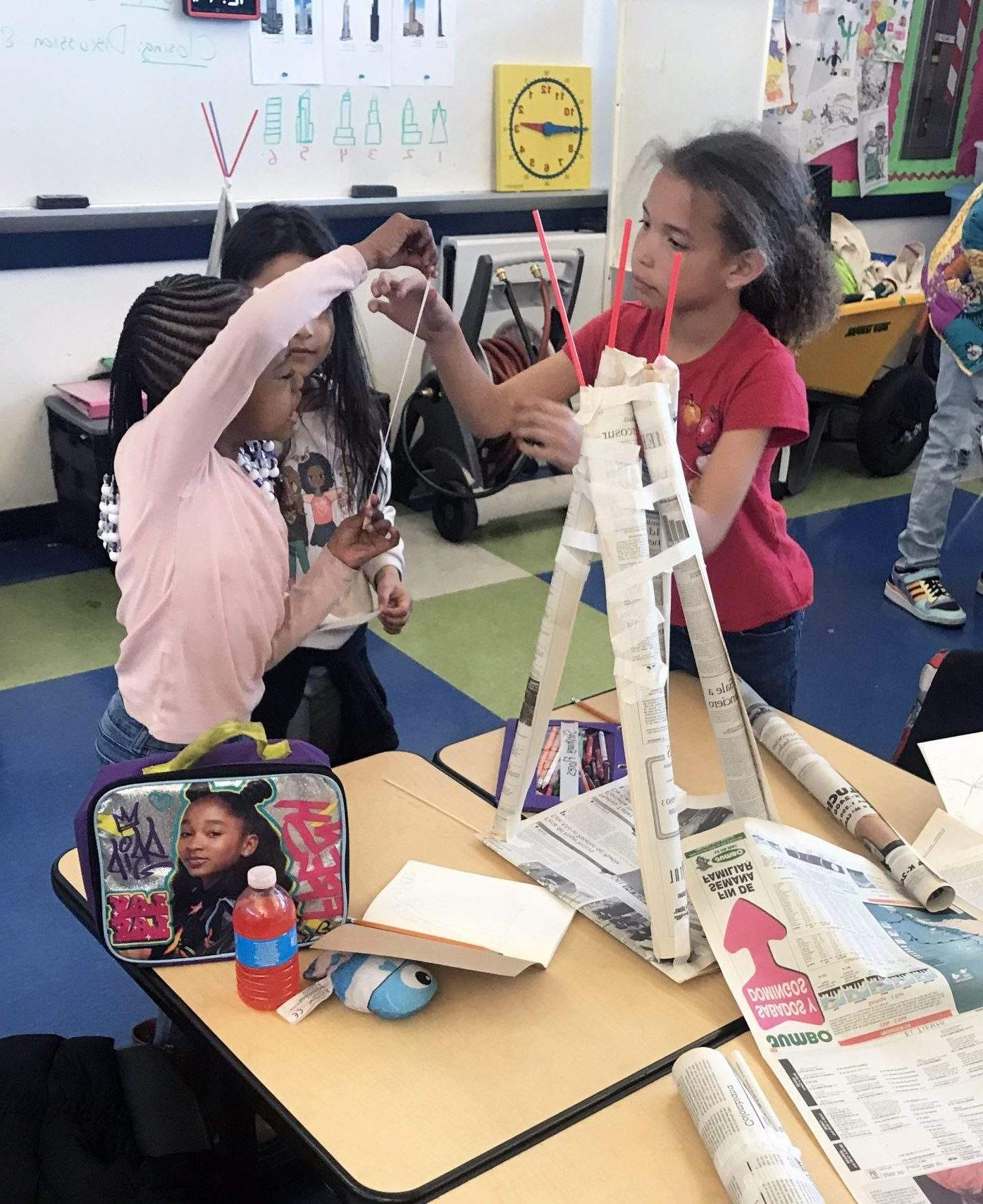 三个女孩准备在背景中添加更多的胶带，用胶带把卷起的报纸粘在一起形成一个棱镜形状的结构.