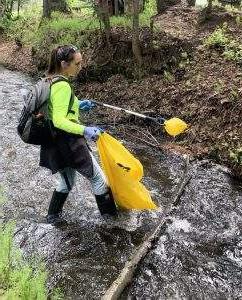 一个穿胶靴的女人, 灰色背包, 工作手套, 一件亮绿色的衬衫站在小溪里，拿着一个抓取器和一个黄色的塑料袋捡垃圾.