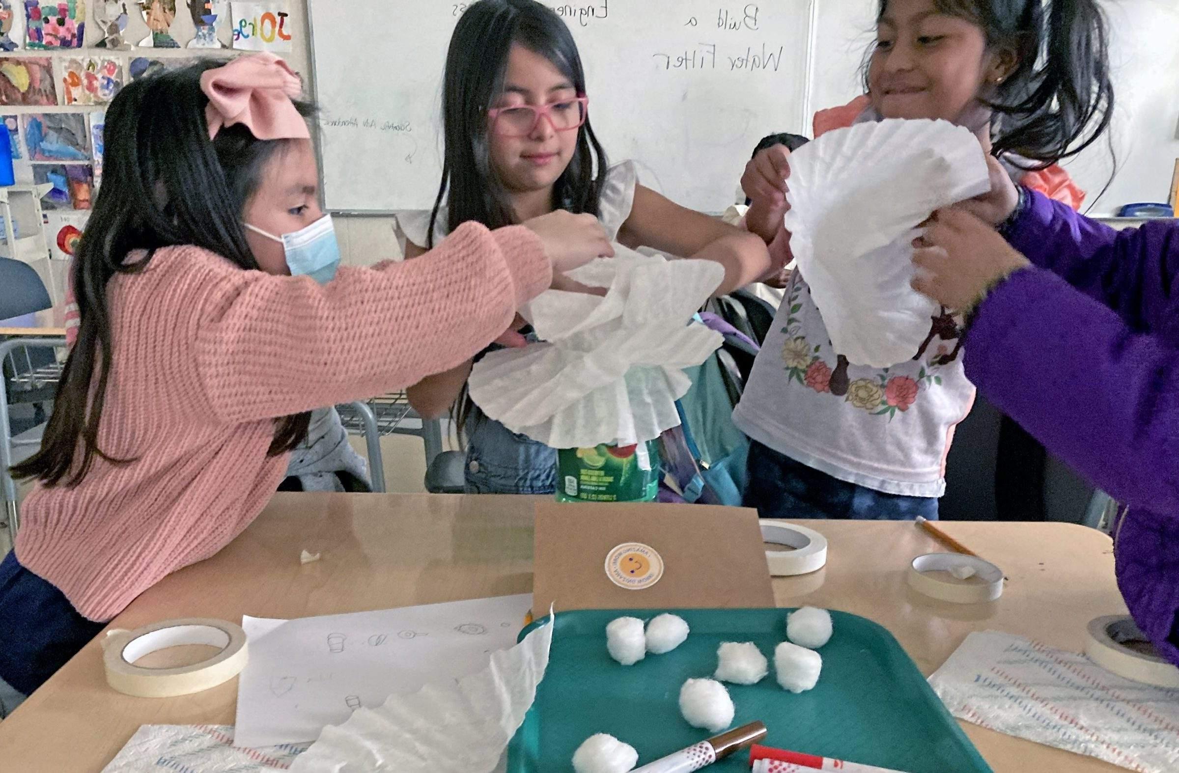女孩们在教室里把咖啡滤纸堆在罐子上. 他们的桌子上放着胶带、棉球、纸巾和记号笔.