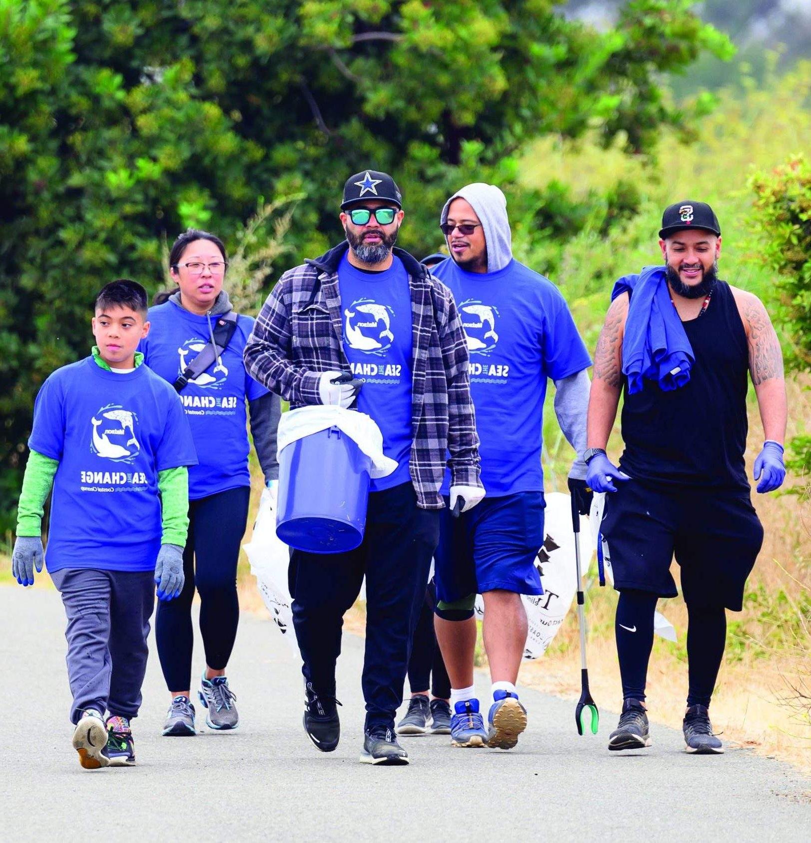 一群志愿者穿着印有“Sea The Change”字样的蓝色澳博体育app下载 t恤，手持抓手, 袋子和水桶, 步行到清理区.
