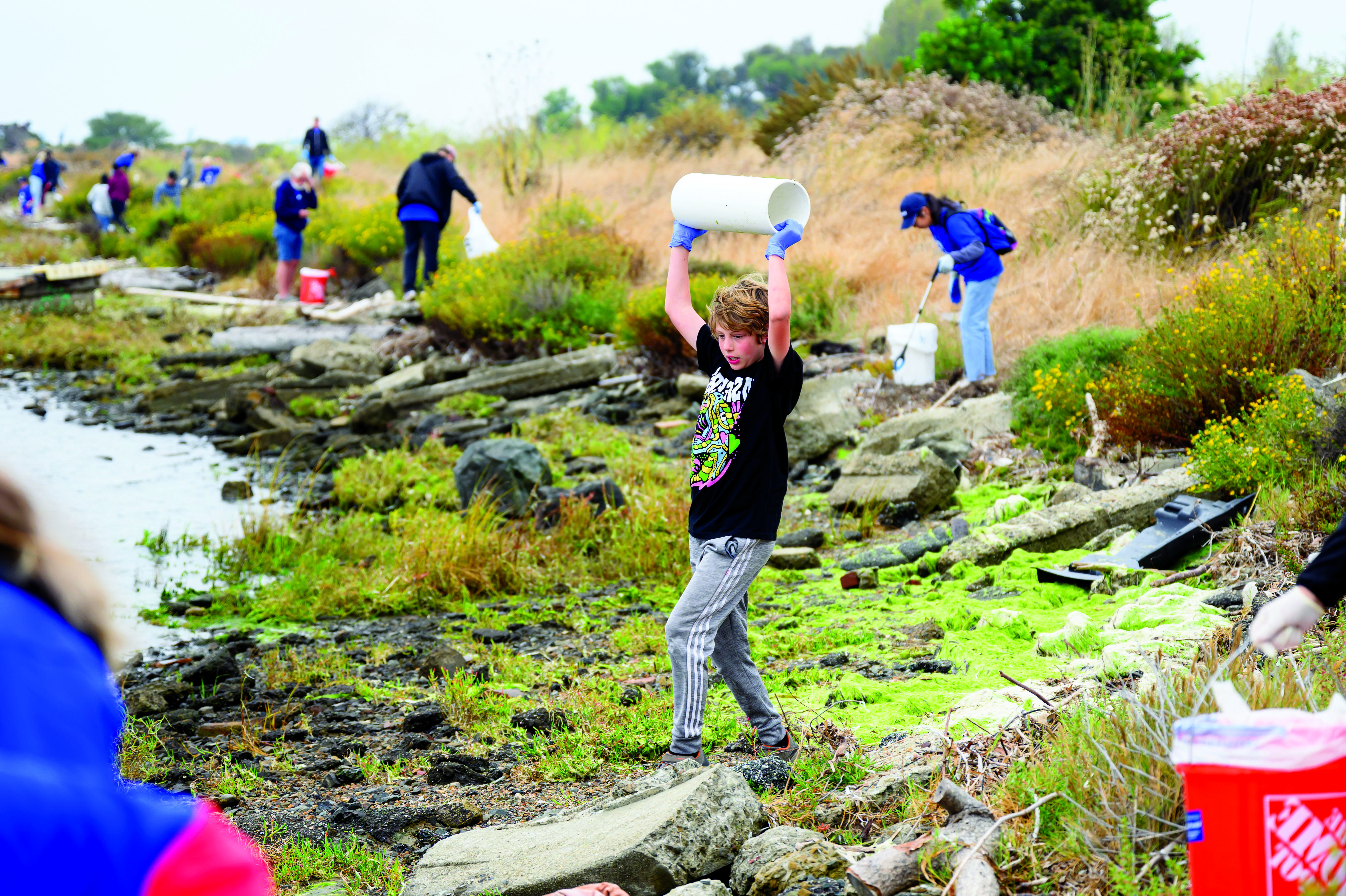 一名戴着蓝色塑料手套的年轻男子在河口拿着一大块塑料管道，背景是其他清理志愿者.