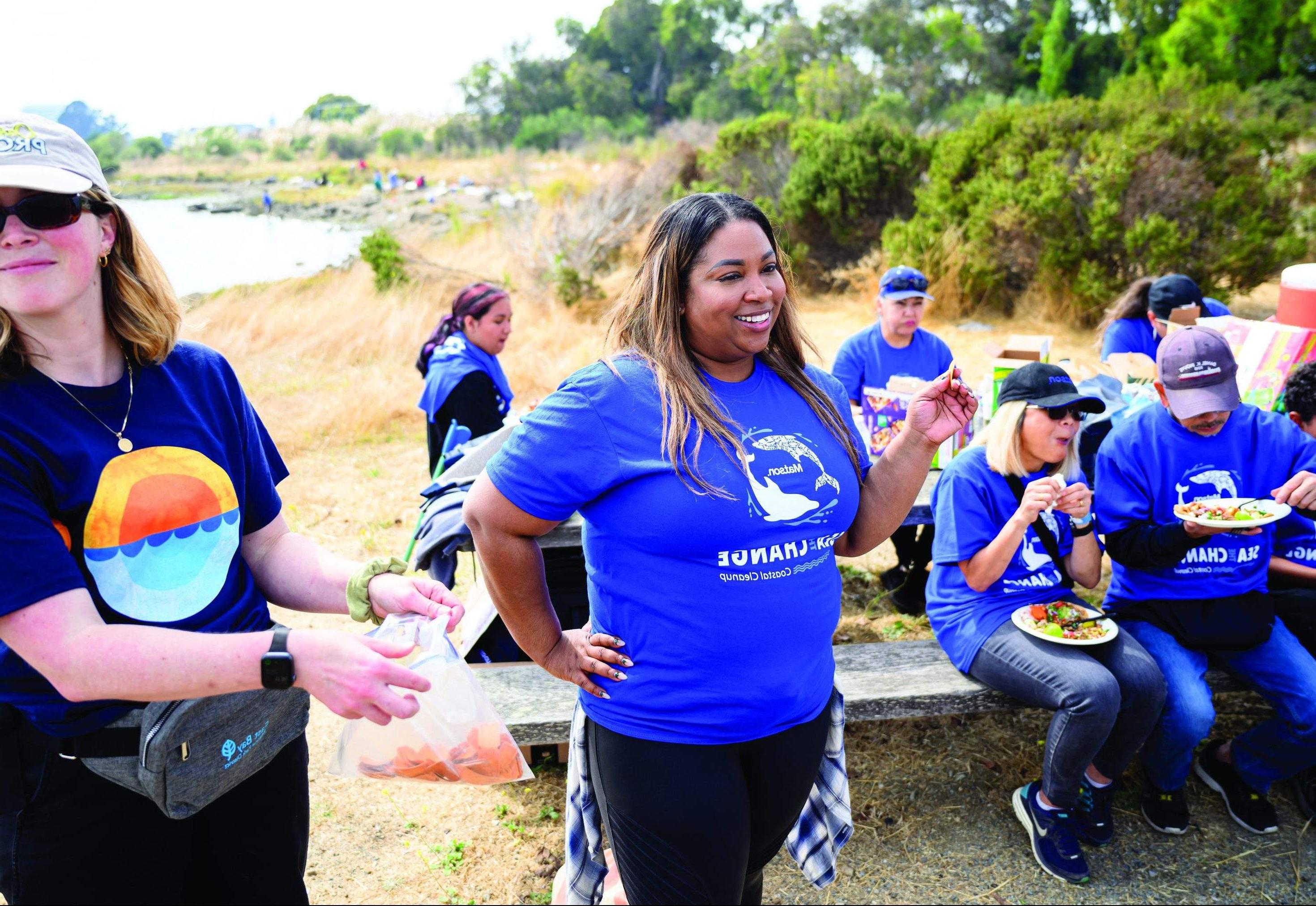 丹尼斯站在那里，手里拿着一张奖券，而志愿者们穿着印有“Sea The Change”字样的蓝色澳博体育app下载 t恤，坐在她身后的野餐桌上吃饭.