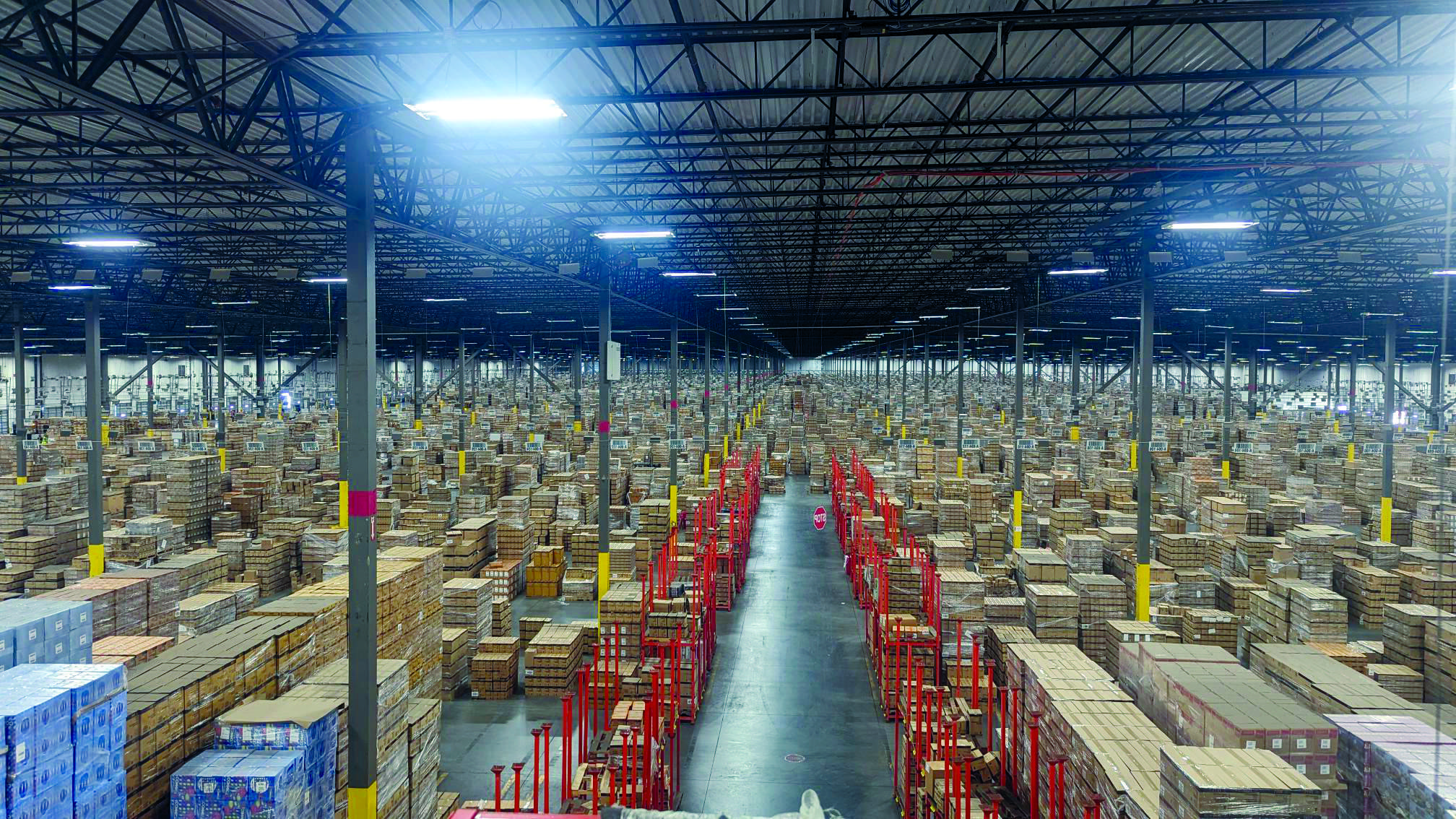Pallets of brown boxes sit stacked in an expansive high ceiling warehouse.