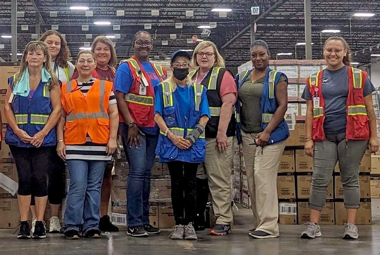 Team members wearing safety vests pose in front of brown boxes.