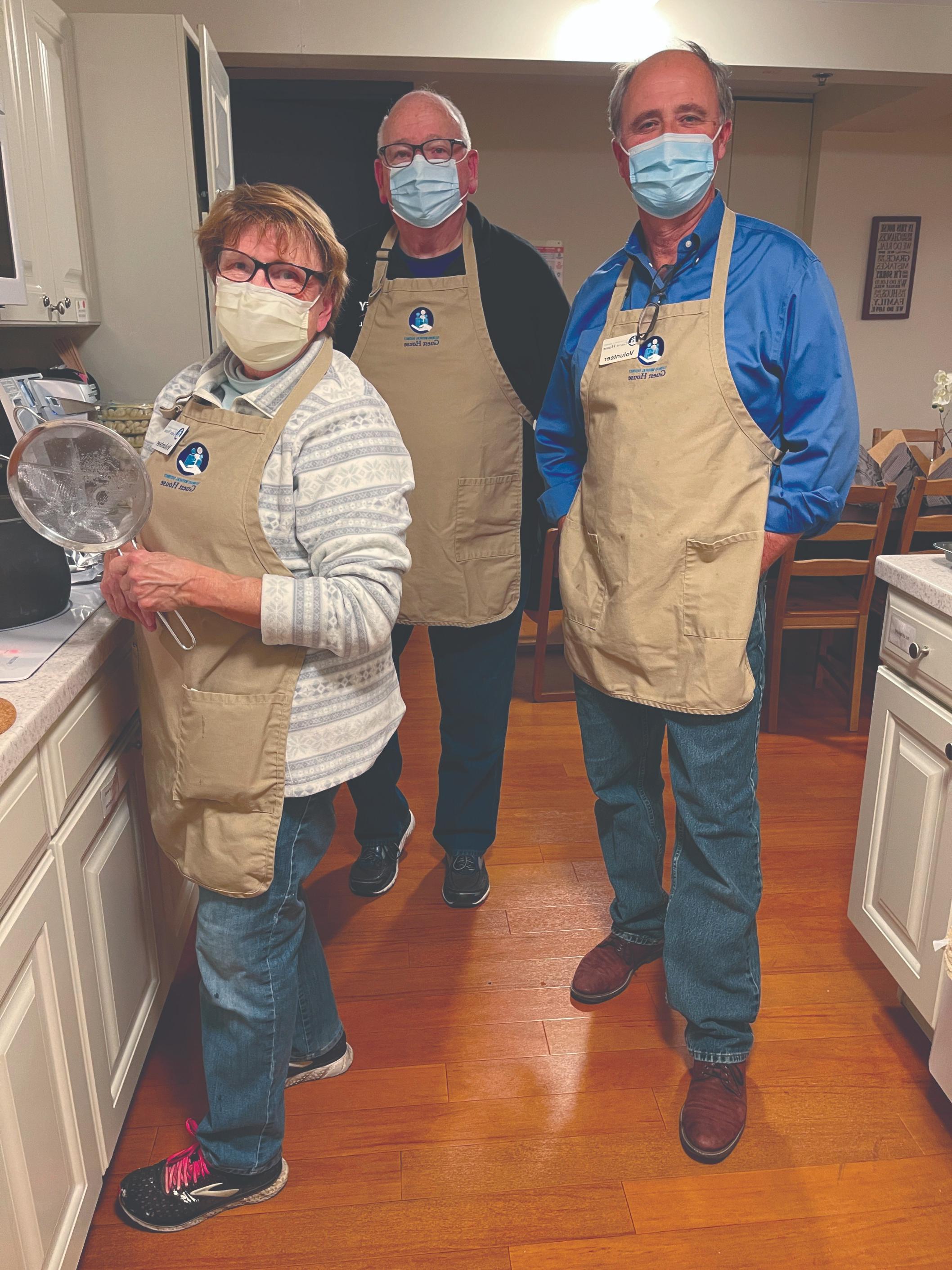 Three voluneers stand in a kitchen wearing masks and beige aprons embroidered with the Guest House logo.