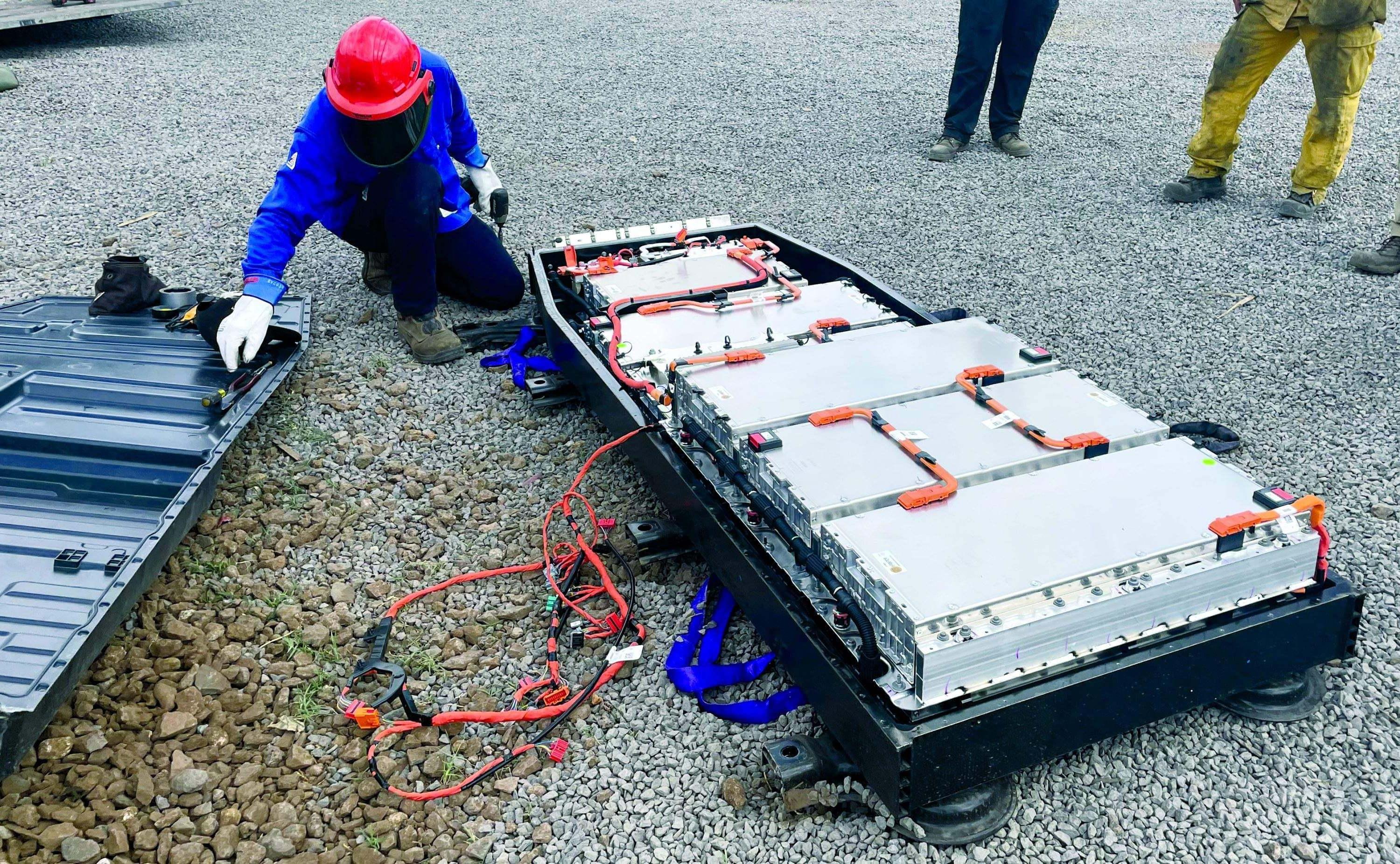 An electrician wearing a red hard hat and protective mask and blue jacket kneels on the ground to disassemble a Ford Lightning battery.