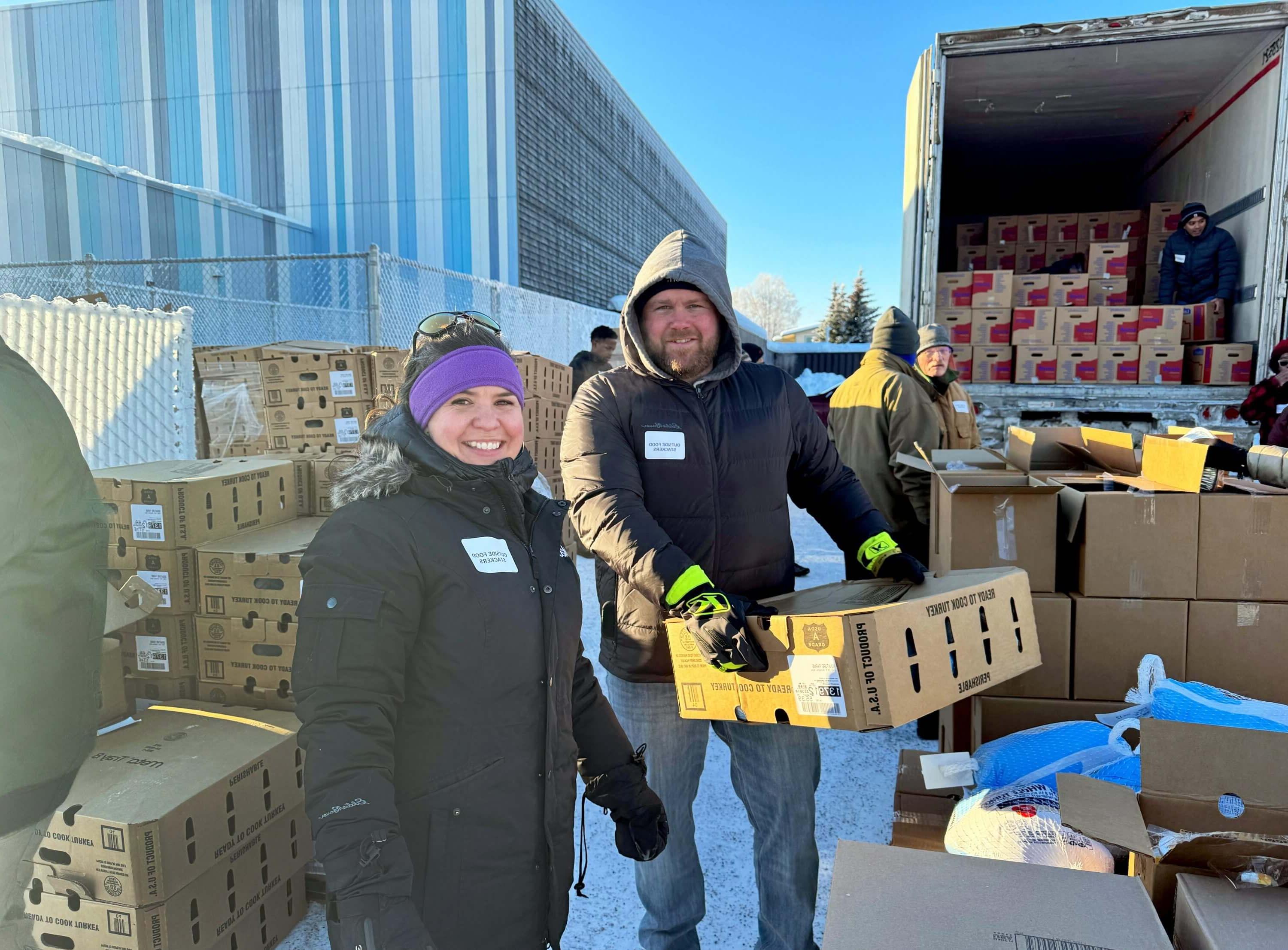 Standing outside and bundled in warm clothing, Joel and Jill Morse help move large boxes of food.